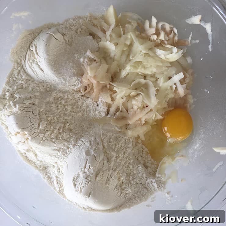 A glass bowl filled with the raw ingredients for Klubb dumpling dough, including grated potatoes, flour, and an egg, waiting to be mixed.