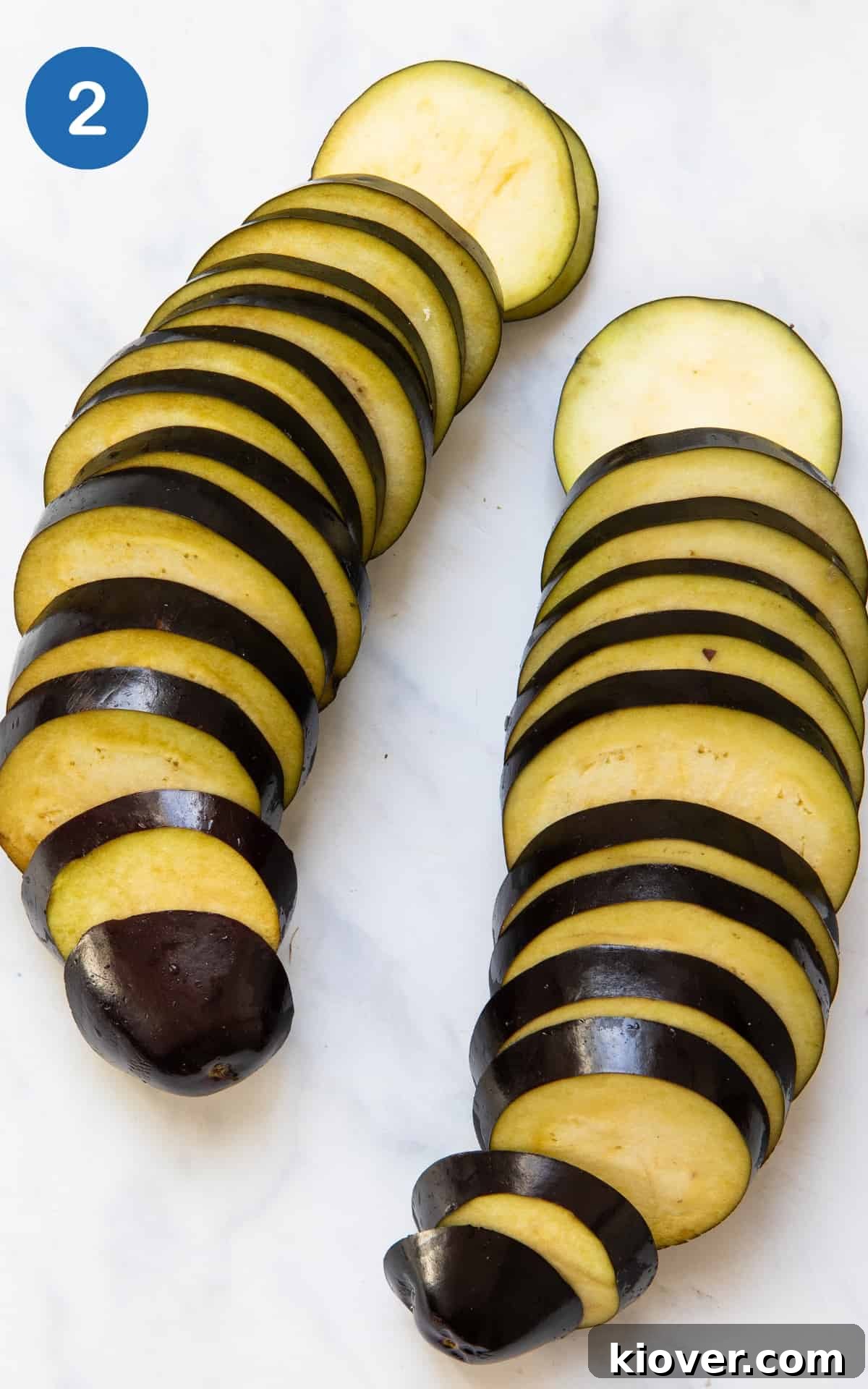 Divine Eggplant Casserole 5 Two aubergines, neatly sliced into uniform circles, are arranged in a straight line on a pristine white marble background, ready for preparation.