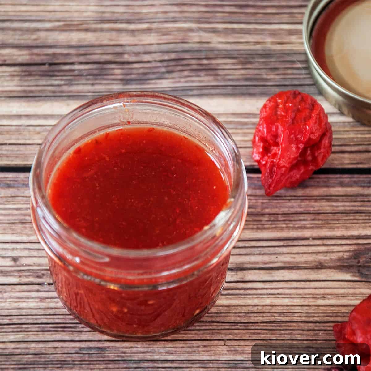A glass jar filled with vibrant red Carolina Reaper Hot Sauce, with a fresh Carolina Reaper pepper resting beside it on a rustic surface.