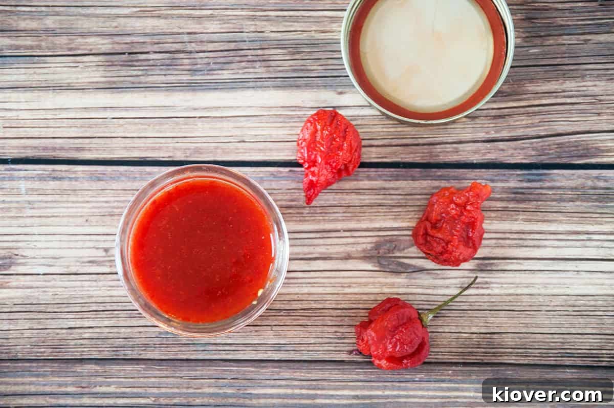 Overhead shot of a jar of homemade Carolina Reaper Hot Sauce, accompanied by three whole Carolina Reaper peppers on a textured wooden table.
