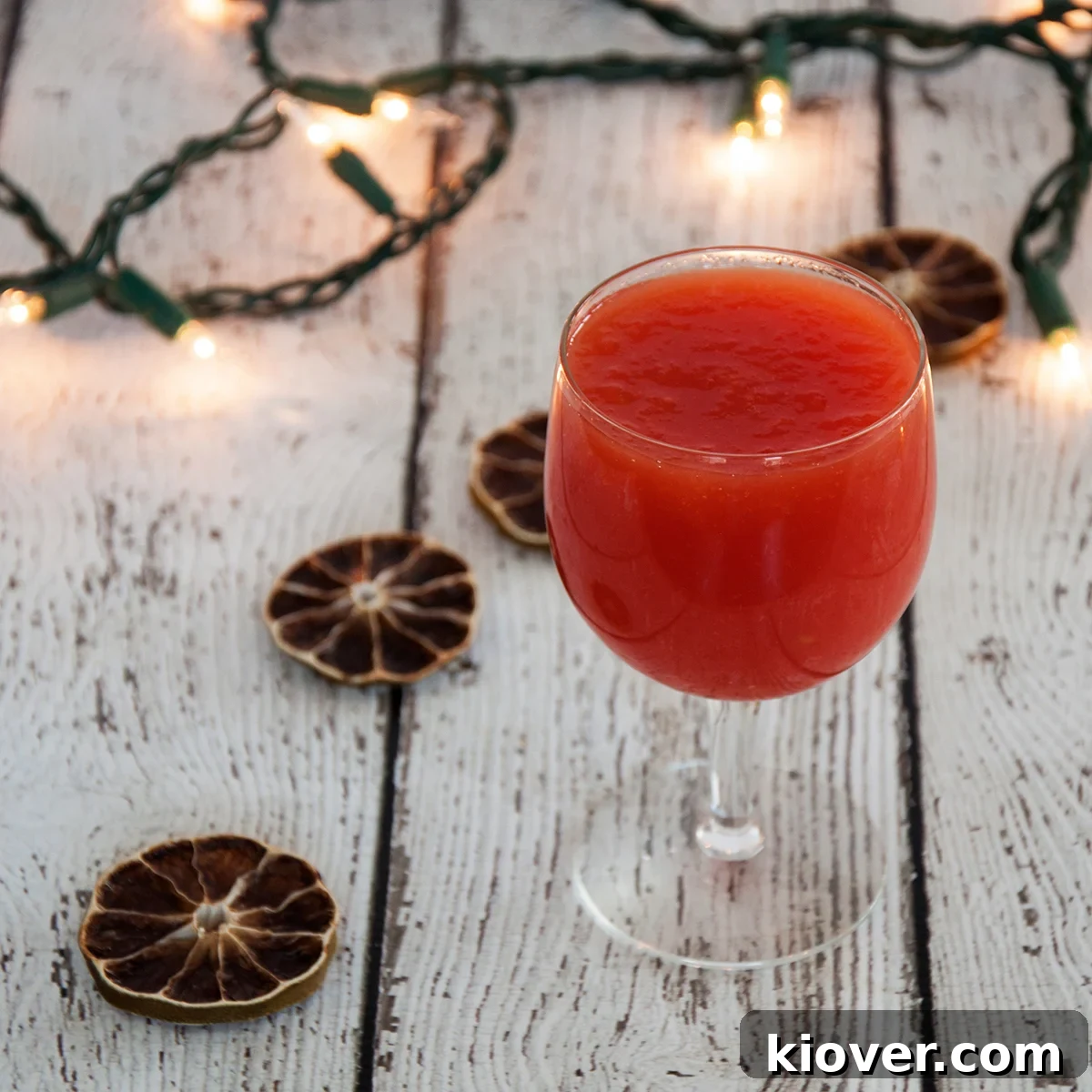 A glass of homemade tomato juice on a wood table with dried limes and holiday lights in the background, signifying a fresh, healthy drink.