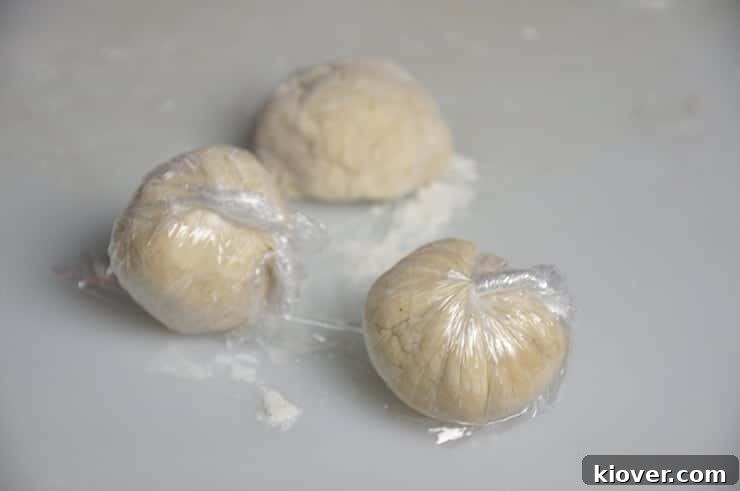 Three perfectly formed dough balls, individually wrapped in plastic wrap, resting before rolling.