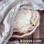 Homemade flour tortillas on a white and red plate with a white towel covering