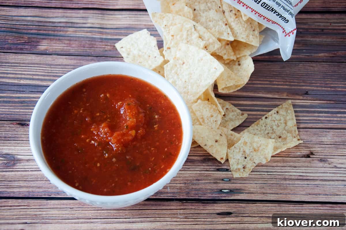 A bowl of salsa with some corn tortilla chips right next to it on a wood table.