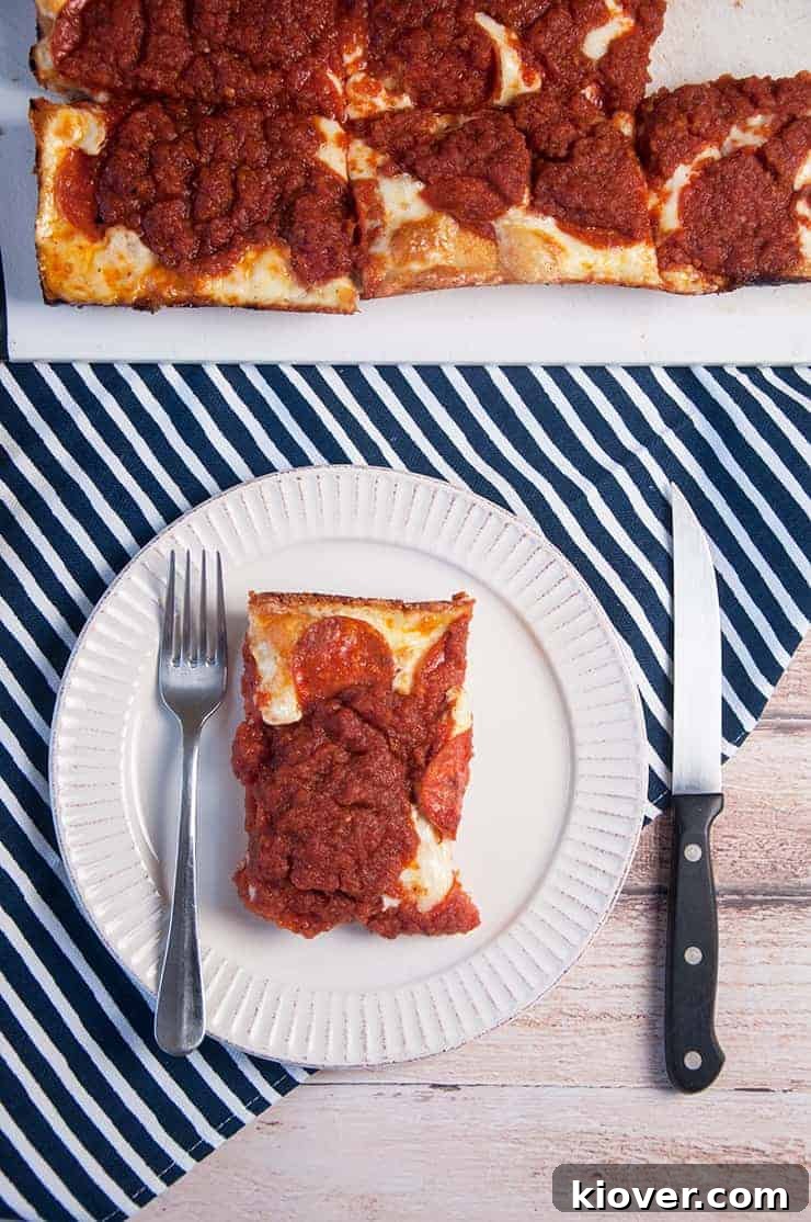 A single, perfectly cut slice of Detroit Style Pizza on a white plate, showcasing its thick, airy crust, generous toppings, and caramelized cheese edge. The full pizza rests on a cutting board in the background.