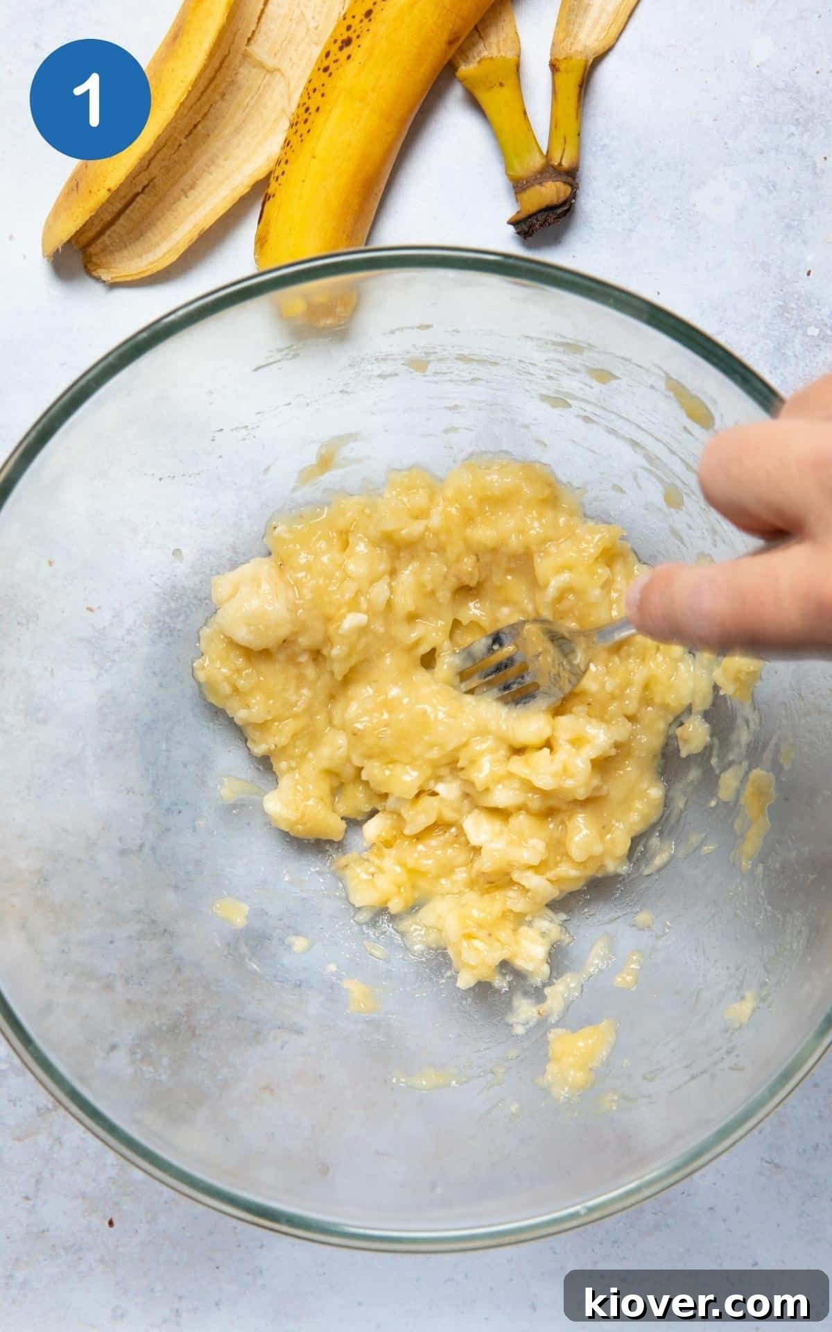 Ripe bananas being mashed with a fork in a large mixing bowl, set against a pristine white background.