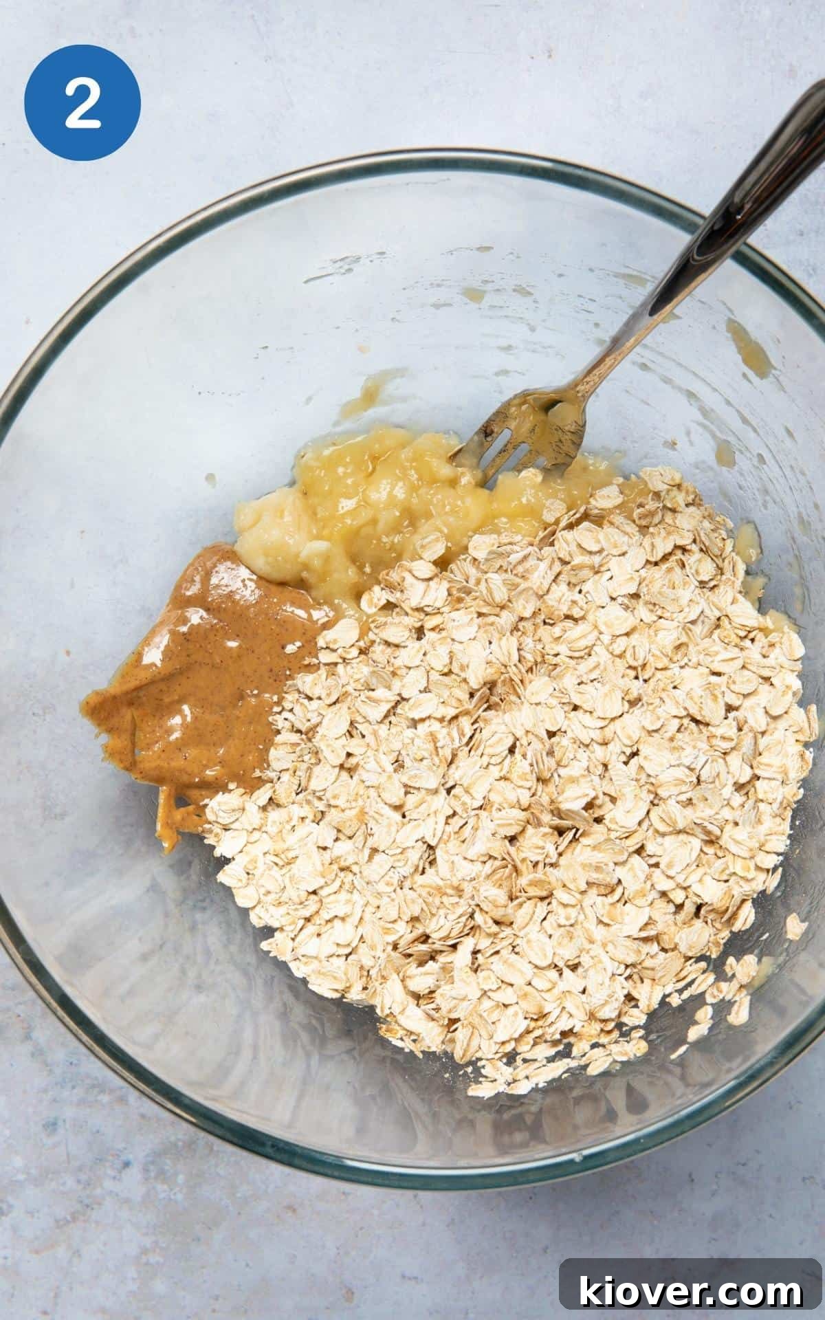 Oats and a dollop of peanut butter being added to the mashed bananas in a large mixing bowl, ready for combining.