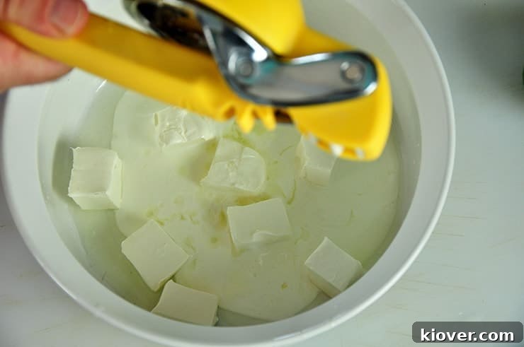 A hand squeezing fresh lime juice into a white bowl containing a mixture of Crema Mexicana and softened cream cheese, forming the base of the creamy sauce.