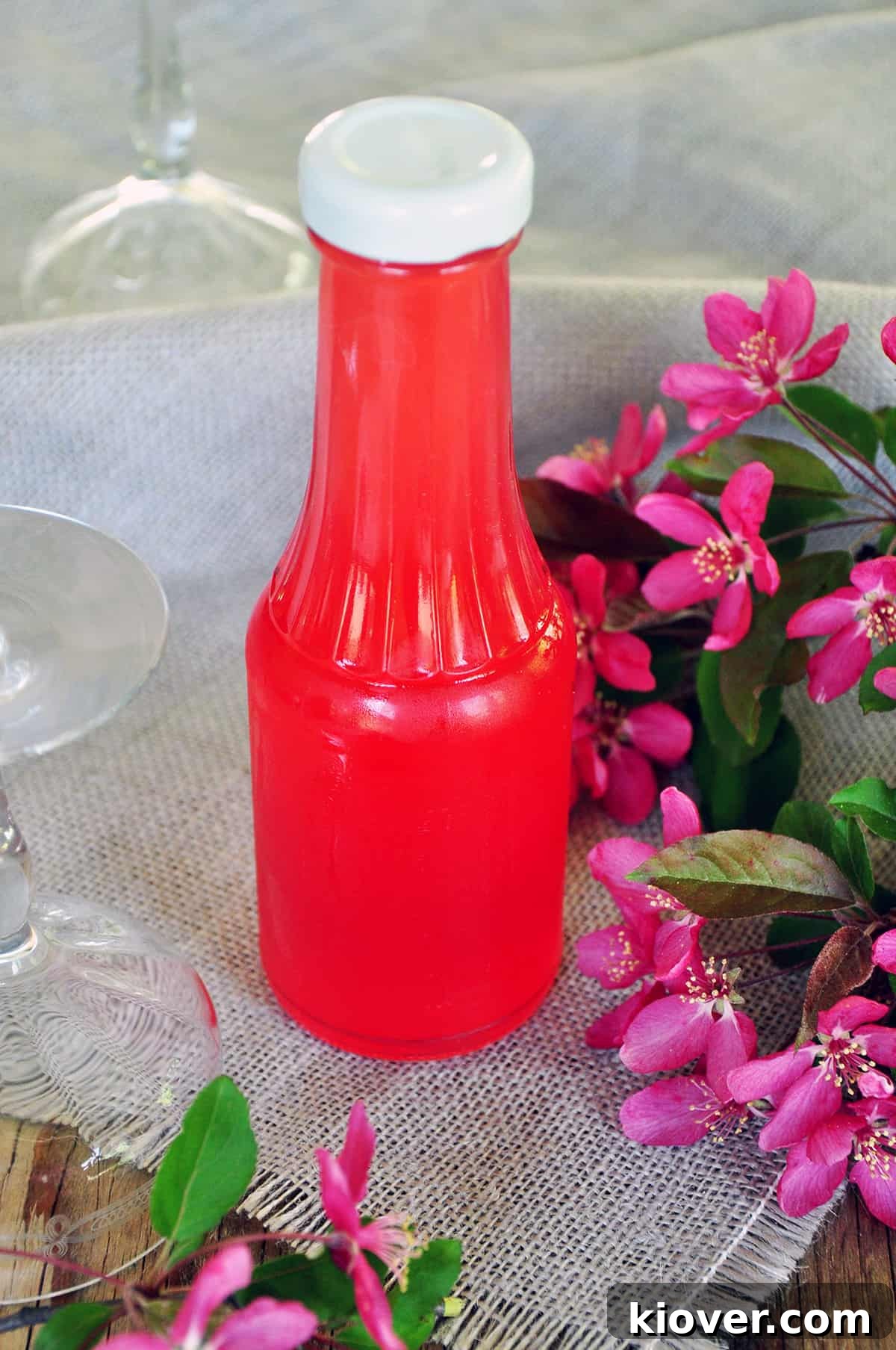 A bottle of homemade Rhubarb Syrup and an empty coupe glass on a picnic table, adorned with pink flowers, hinting at a refreshing cocktail moment.