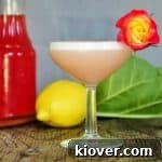 Limoncello and Rhubarb Cocktail on a pice of wood with rhubarb, lemon, and syrup in the background. The cocktail is garnished with a red and yellow rose.