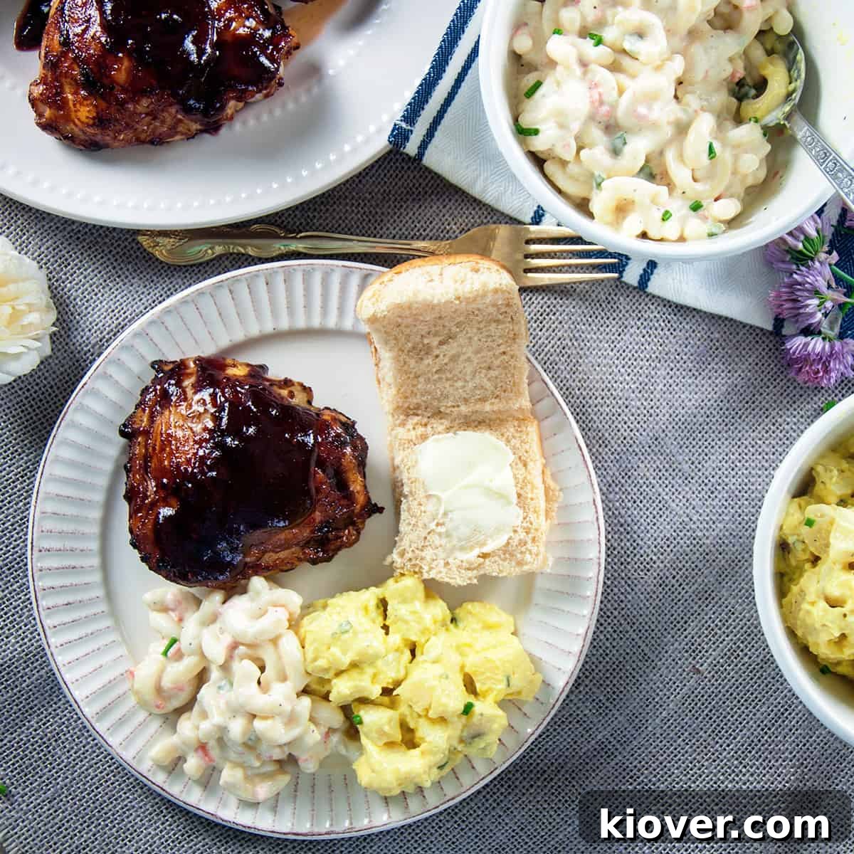 Grilled BBQ Chicken Thigh on a plate with potato salad, macaroni salad, and a Hawaiian Bun.