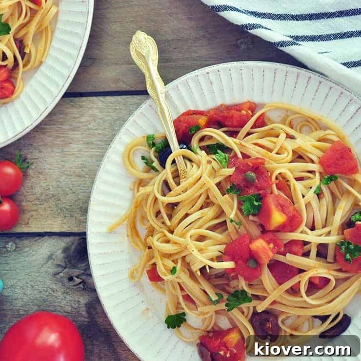 A plate of Puttanesca Sauce with Linguine on a white plate with a gold spoon. It is in a picnic table with a striped white cloth and some whole tomatoes, ready to be enjoyed.