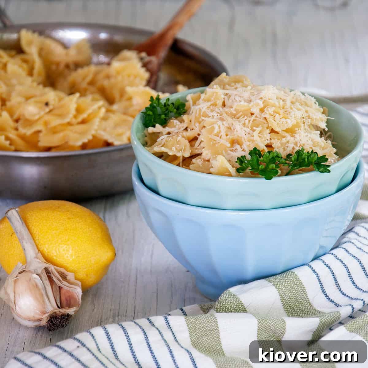 A bowl of Creamy Lemon Pasta Sauce with Farfalle noodles, with the rest of the skillet and ingredients like a napkin, lemon, and garlic in the background on the table.