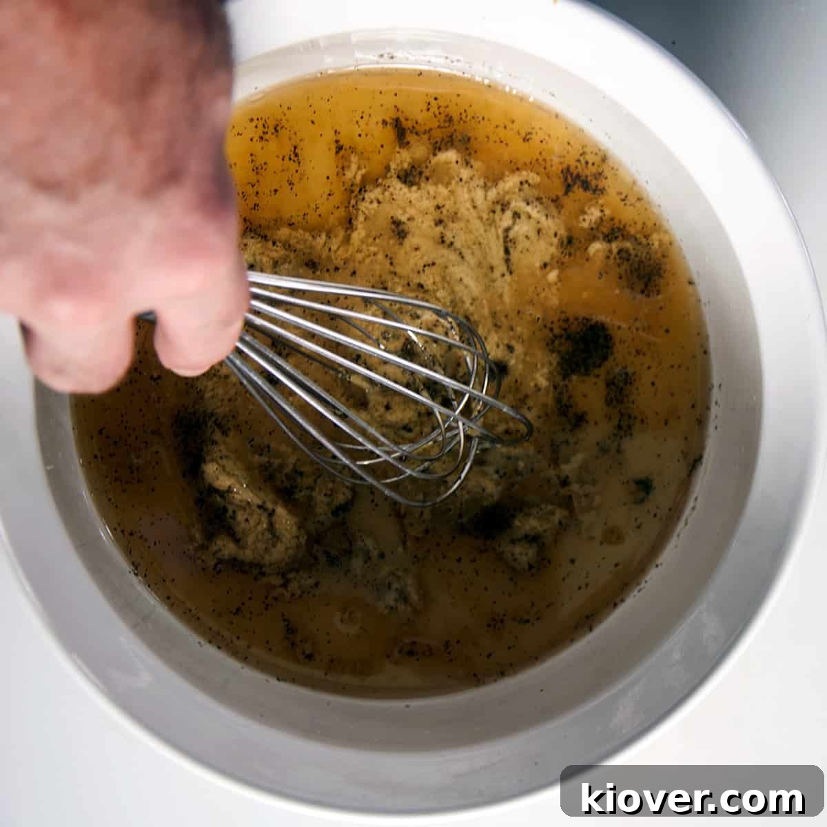 A whisk vigorously mixing wet dressing ingredients including vinegar, mustard, and oil in a clean white bowl for the Kartoffelsalat.