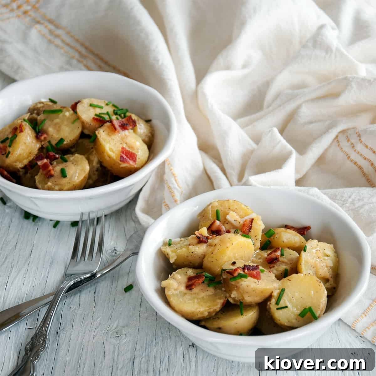 Two enticing bowls of traditional warm German potato salad (Kartoffelsalat) generously garnished with fresh green chives and golden-brown crispy bacon, set against a clean white background.