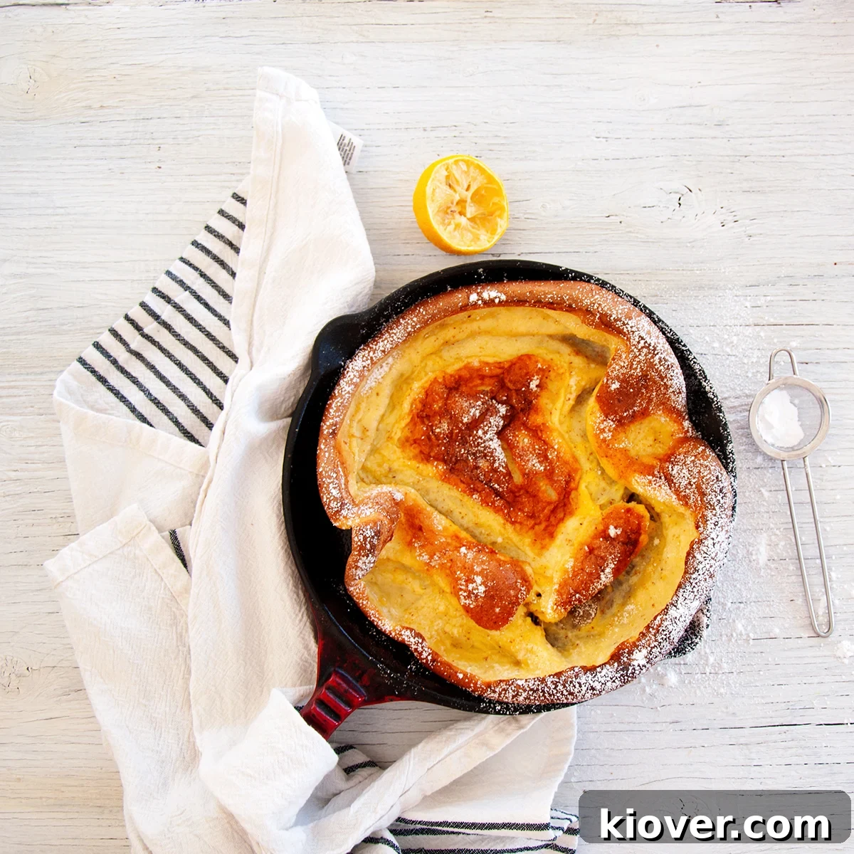 Overhead view of a perfectly puffed, golden Dutch Baby Pancake, dusted with powdered sugar and drizzled with lemon juice, served in a cast-iron skillet.