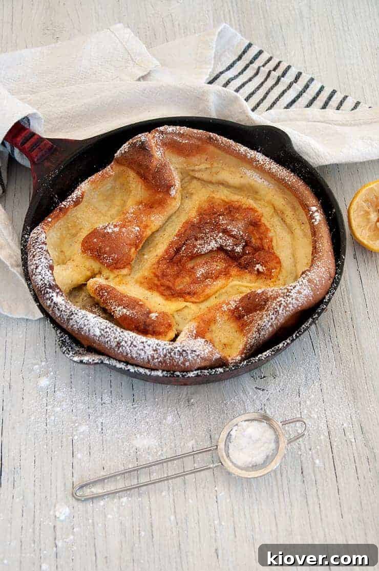 Side view of a perfectly risen Dutch Baby Pancake, golden brown and fluffy, resting on a white table within its cast-iron skillet.