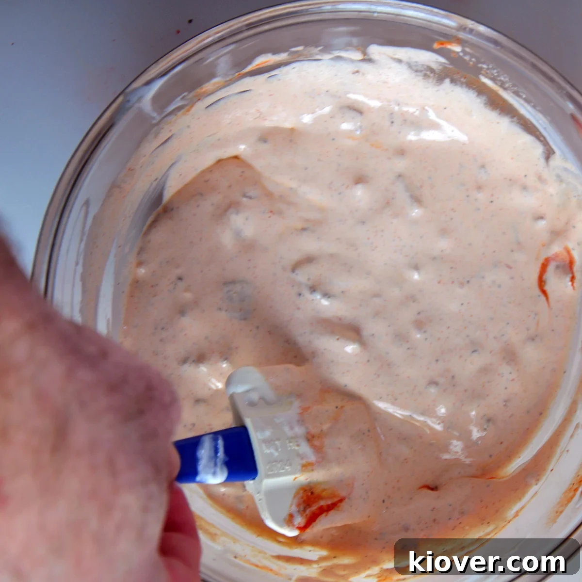 Mixing away A close-up shot of a spoon actively mixing the sriracha chip dip in a bowl. The creamy texture is visibly swirling, blending the vibrant red sriracha and golden bacon bits into the sour cream base, indicating a thorough combination of ingredients.