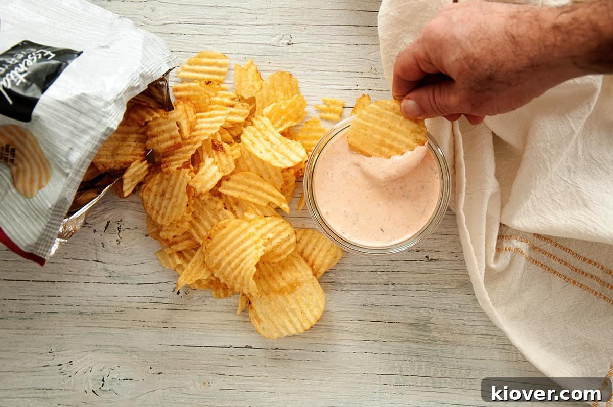Bacon and Sriracha Chip Dip An inviting overhead shot shows a hand reaching into a mason jar of Sriracha and Bacon Chip Dip with a single potato chip, poised to scoop up a creamy, flavorful bite. Golden potato chips are scattered around the jar on a white surface, highlighting the delicious anticipation.