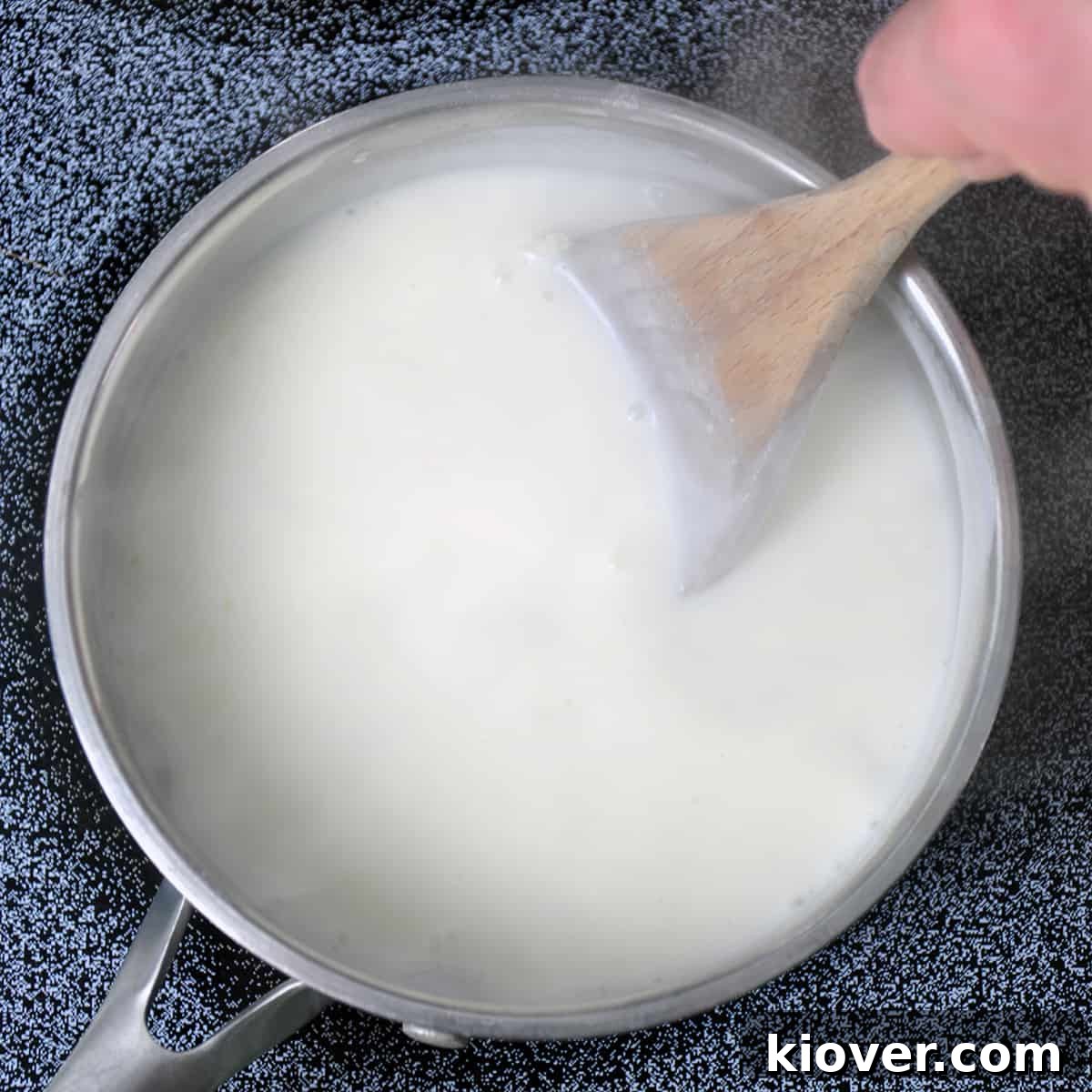 Mornay Sauce Preparation A metal pot with mornay sauce being thickened, showing a whisk stirring the creamy white sauce.