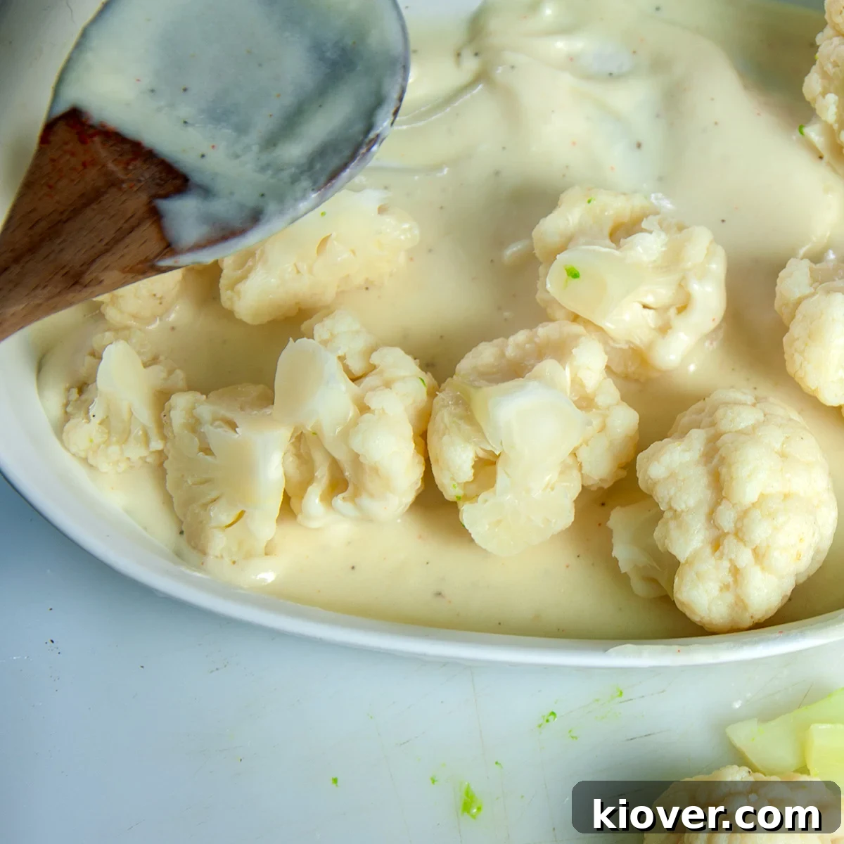 Mixing Cauliflower with Sauce A casserole dish with cooked cauliflower florets being mixed with mornay sauce using a wooden spoon.