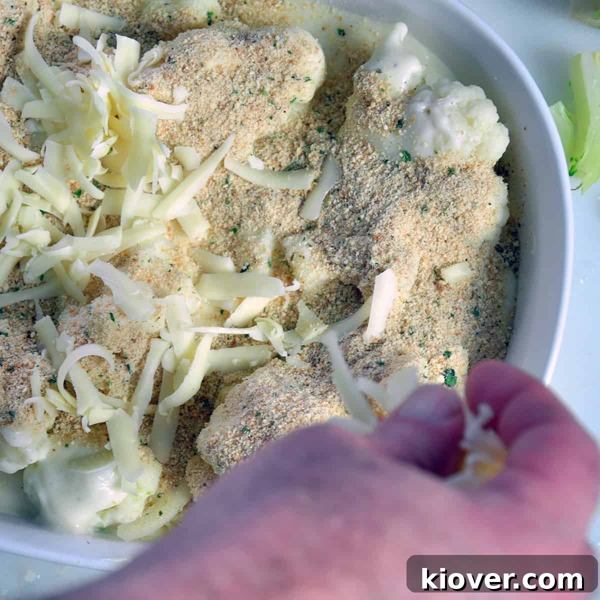 Topping with Cheese and Breadcrumbs A close-up of shredded cheese and breadcrumbs being sprinkled over an unbaked cauliflower au gratin in a casserole dish.