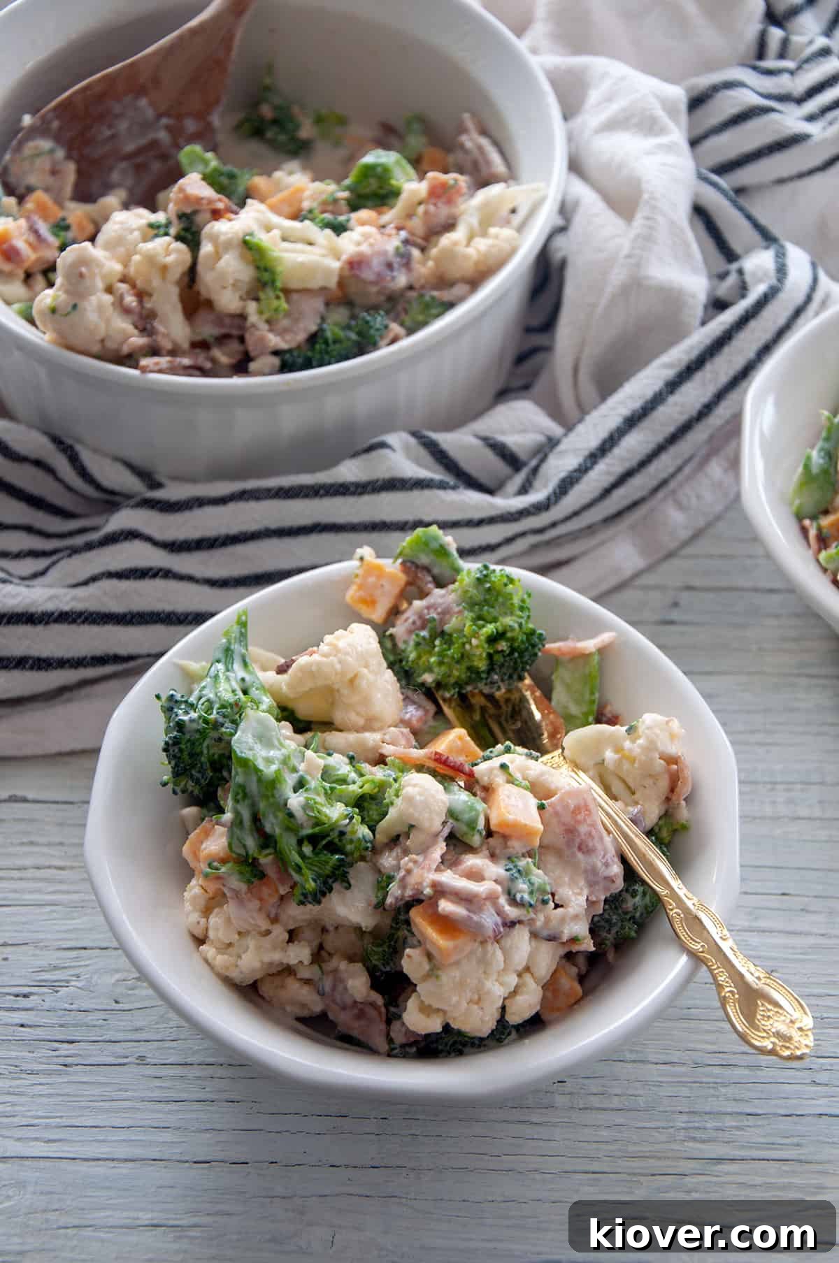 A large white bowl brimming with Extra Baconey Amish Broccoli Salad, garnished with crumbled bacon and fresh herbs.