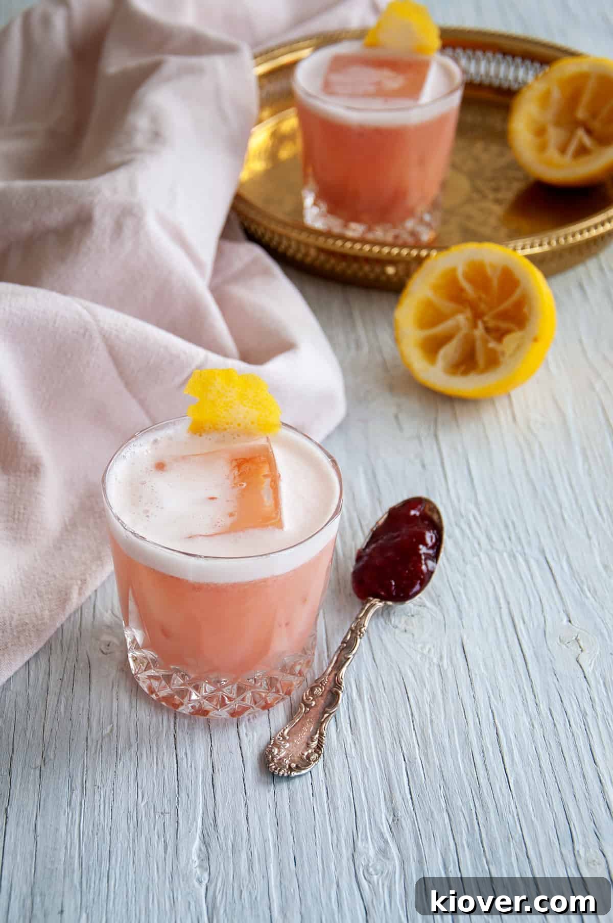 A forty-five-degree angle shot of our beautifully presented Strawberry Jelly and Gin Cocktail, resting on a clean white table, accompanied by an elegant spoon artfully filled with strawberry jelly, highlighting the star ingredient.