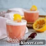 Two Strawberry Jelly and Gin Cocktails on a white table with a spoon of jelly and a juiced lemon on the side.