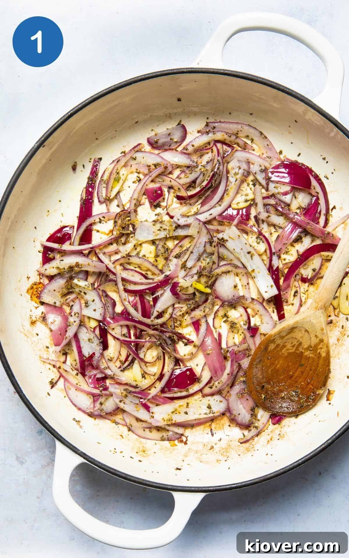 Sliced red onions being gently fried in olive oil in a large non-stick frying pan, with a wooden spoon for stirring. The pan is on a white marble surface.