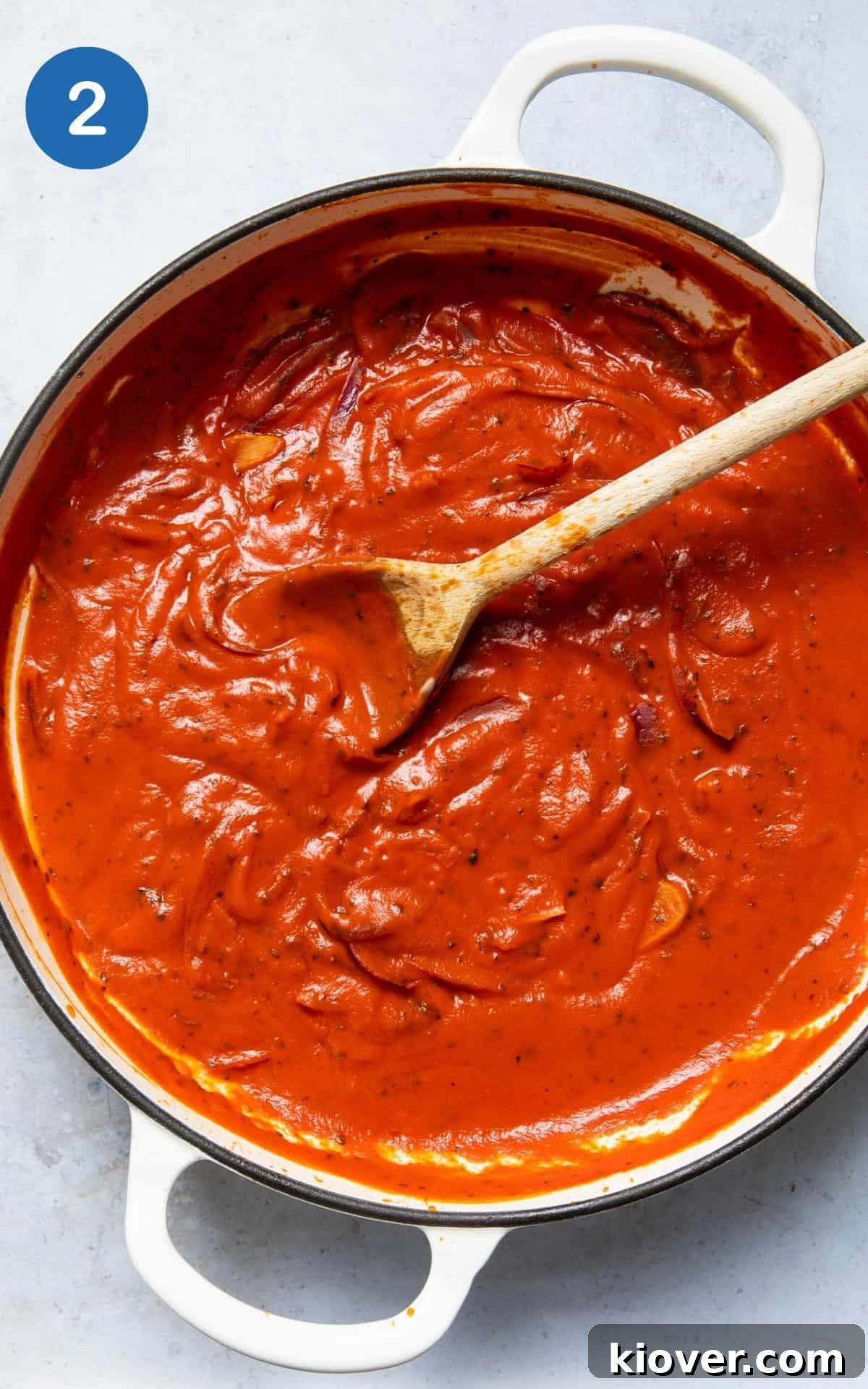 Tomato passata and coconut yogurt being stirred into the softened onions and garlic in a large non-stick frying pan, forming the base of the creamy tomato soup.