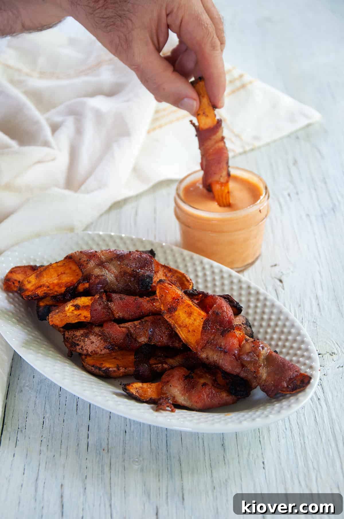 Smoky Bacon-Wrapped Grilled Sweet Potato Sticks 6 A hand reaches into a mason jar of Sriracha Maple dipping sauce, pulling out a perfectly grilled, bacon-wrapped sweet potato fry. The white plate beside it is filled with more delicious fries, ready to be devoured.