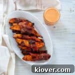 A crisp feature image of bacon-wrapped grilled sweet potato fries on a white plate, accompanied by a mason jar of Sriracha Maple dipping sauce.
