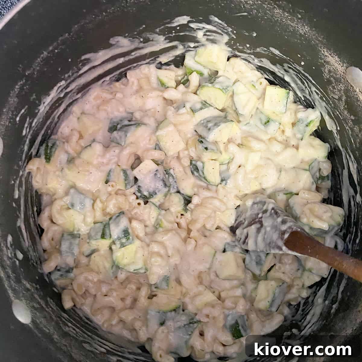 Dish where zucchini, macaroni, and cheese sauce are being mixed with a wooden spoon.