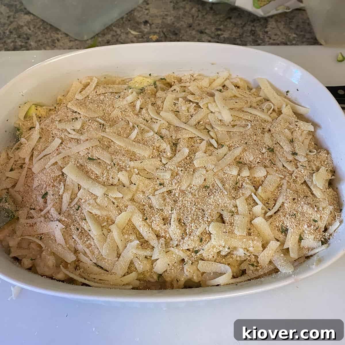 White casserole dish with pre-baked ingredients, including zucchini casserole, cheese, and bread crumbs