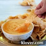 Homemade Queso Dip in a white and gold bowl and a hand dipping a single corn chip into it.