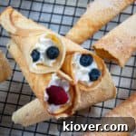 Three Krumkake filled with whipped cream, blueberries, and raspberries that are laying on top of several unfilled ones that have been sprinkled with powdered sugar.