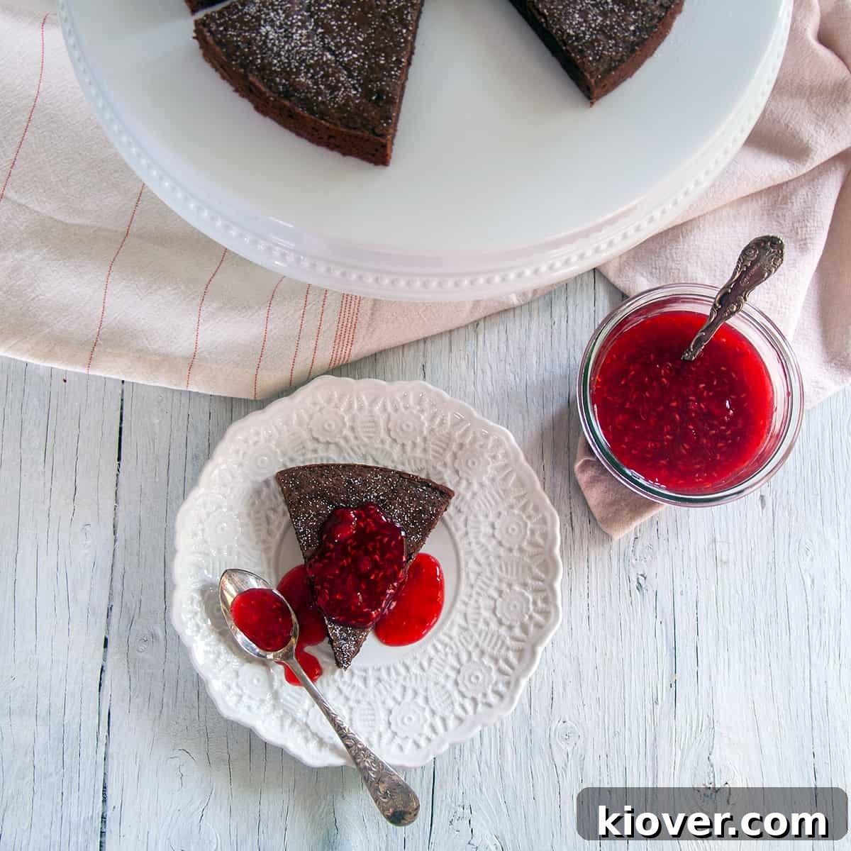 A delectable slice of Kladdkaka Swedish Sticky Cake, generously topped with fresh raspberry sauce and served on a pristine white plate, accompanied by a spoon for the perfect bite.