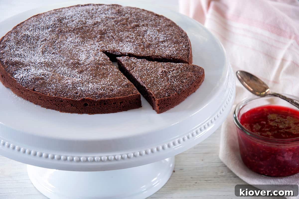 The entire Kladdkaka Swedish Sticky Cake presented on a pristine white serving platter, with a single slice elegantly cut out to reveal its gooey interior, and a bowl of vibrant raspberry topping positioned invitingly to the side.