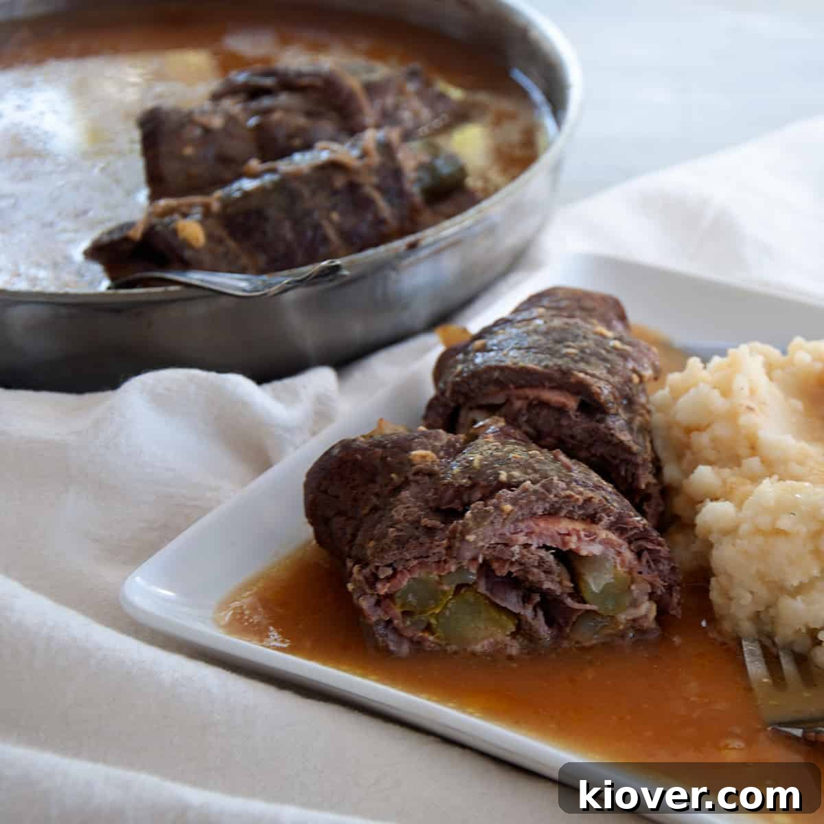Rouladen cut in half to reveal bacon and pickles, but is laying on a white plate with mashed potatoes and gravy.