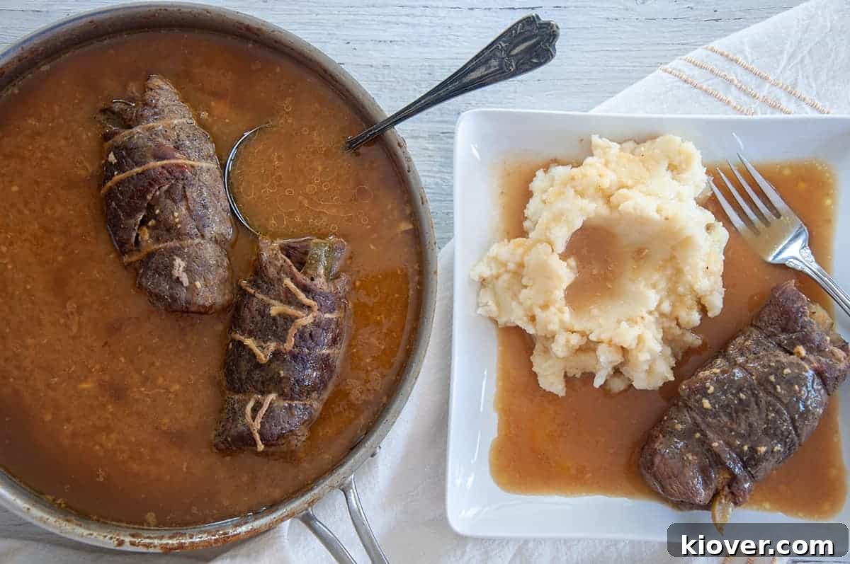 One rouladen on on a white plate with gravy and mashed potatoes, while two others are still sitting in the simmering pan.