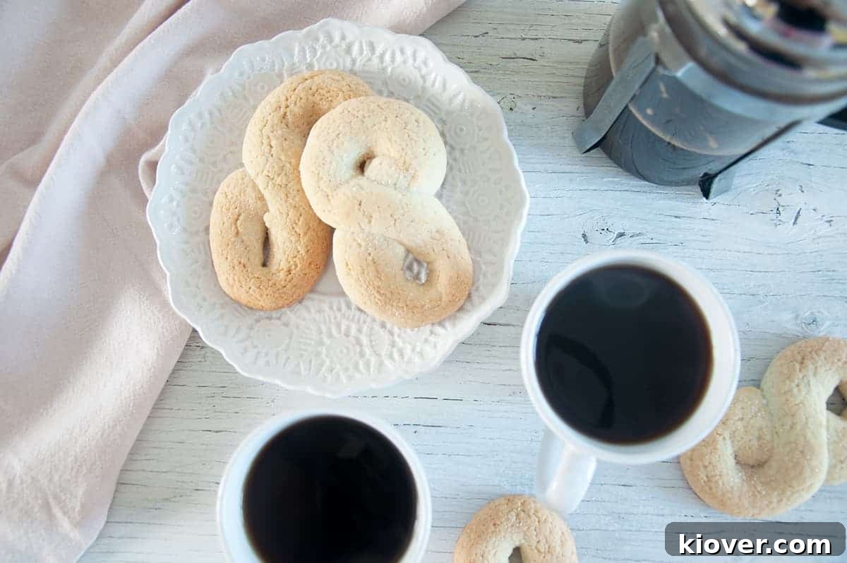 Two kringler on a white plate with two cups of coffee and a French press off to the side.