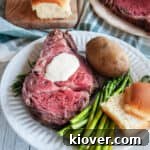 Slice of Prime Rib on a white plate being served with asparagus, a bun, a baked potato, and the entire roast barely showing in the background.