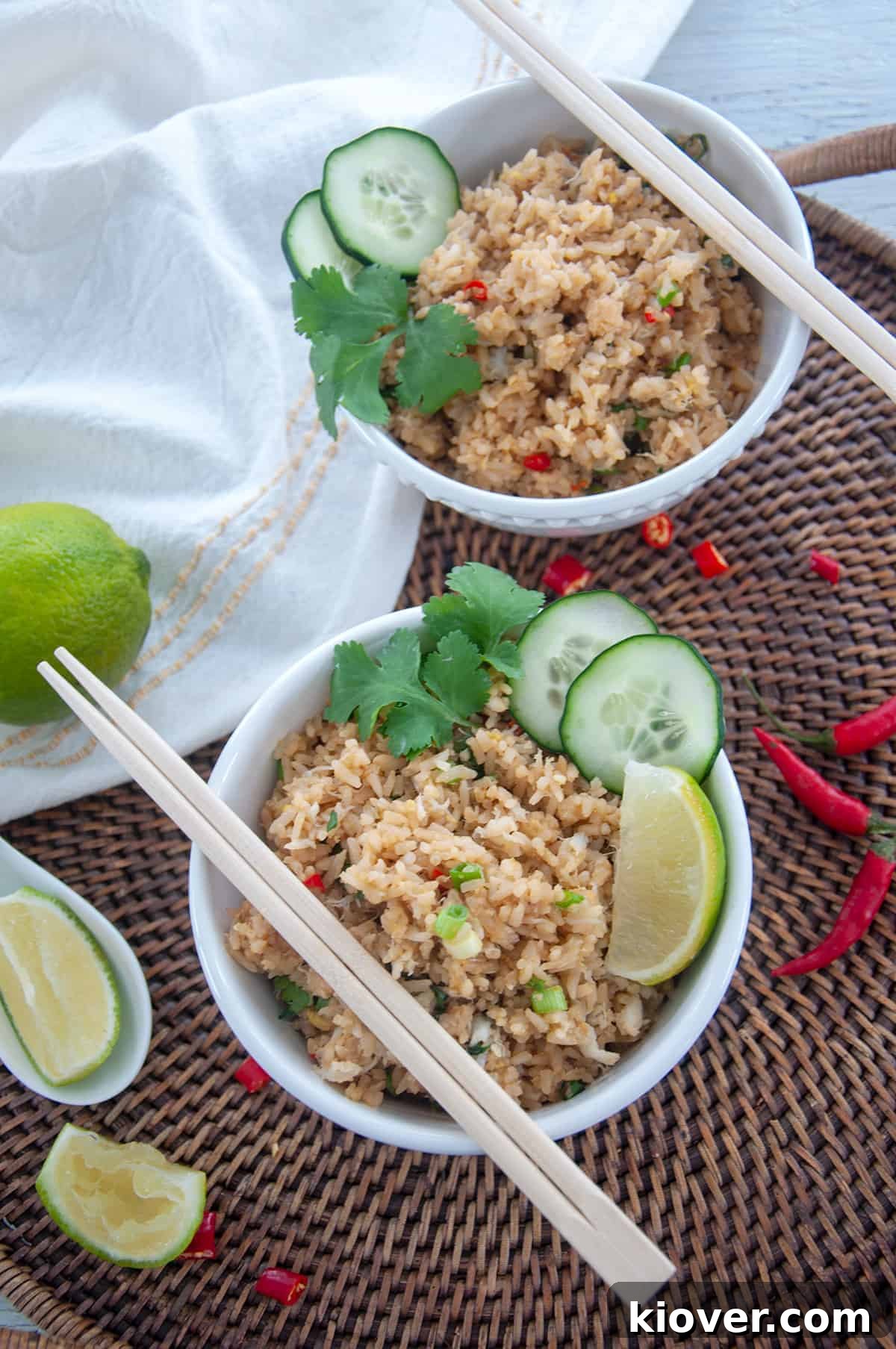 Crab Fried Rice in two white bowls with lime and green onions off to the side. This bowl is sitting on a wicker tray that has a spent lime wedge, thai chili peppers, and a whole lime.