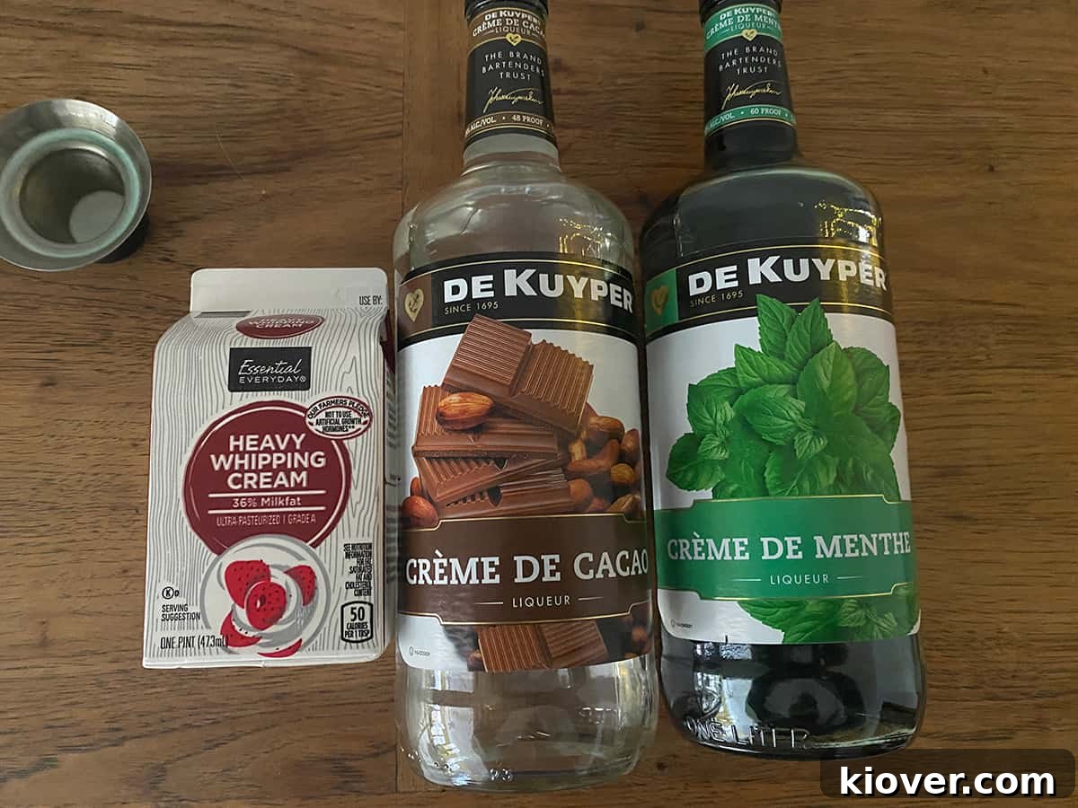 A visual display of the key ingredients for a Grasshopper cocktail: a carton of heavy whipping cream, a bottle of Crème de Cacao, and a bottle of Crème de Menthe, arranged on a rustic wooden table.