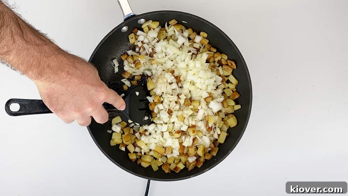 Crispy Homemade Corned Beef Hash 4 A hand expertly stirs a medley of potatoes and onions in a skillet, ensuring even cooking and caramelization for the corned beef hash.