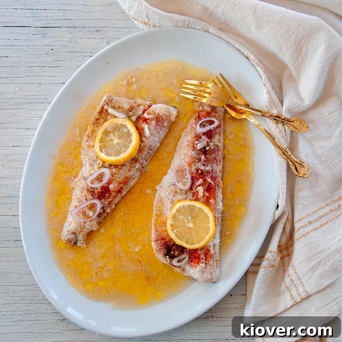 One baked walleye fillet with a lemon slice on top and sitting in white wine sauce and gold forks to the side.