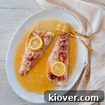 One baked walleye fillet with a lemon slice on top and sitting in white wine sauce and gold forks to the side.