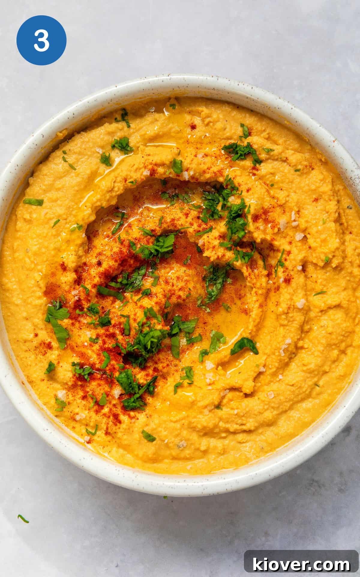 Creamy Garlic Hummus Recipe for Smooth, Flavorful Dip 6 garlic hummus served in a white bowl on a marble worktop topped with paprika, olive oil and fresh parsley.