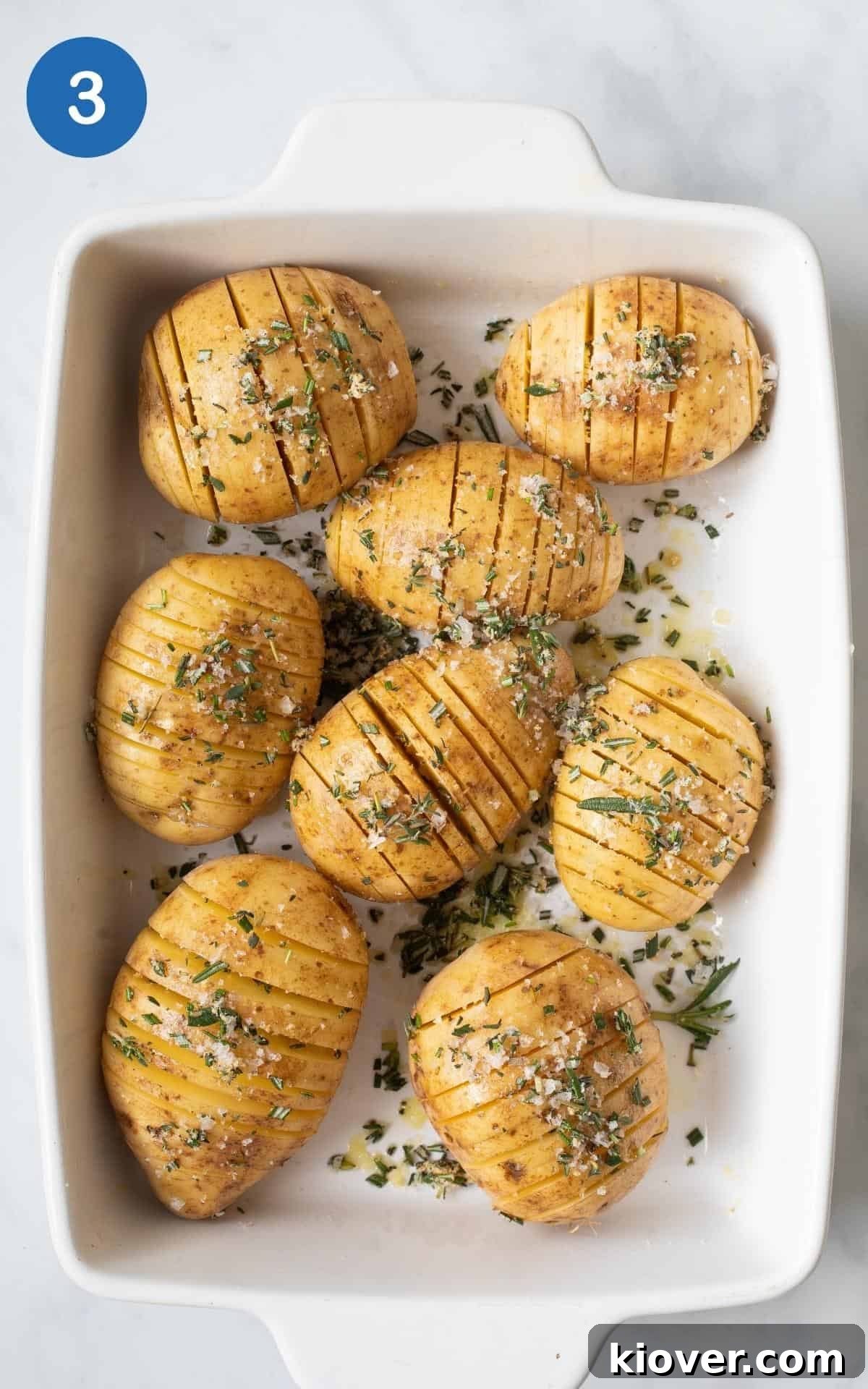 Crispy Hasselback Potatoes with Garlic and Parmesan 6 Sliced potatoes on a baking plate with thyme, rosemary, salt, garlic granules and olive oil on top.